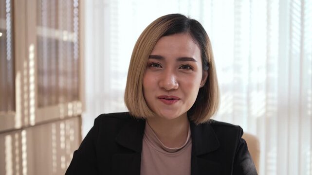 Happy Asian businesswoman talking in a video call at an office desk. Concept of remote work, online meeting, webinar, and digital communication. Professional female looking at camera smiling.
