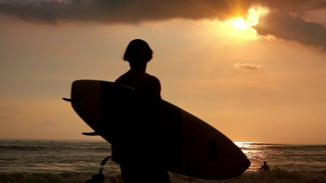 Silhouettes of surfer carrying surfboard and man running along shoreline in evening against ocean illuminated by setting sun on background. People on beach washed by sea at sunset. Slow motion.