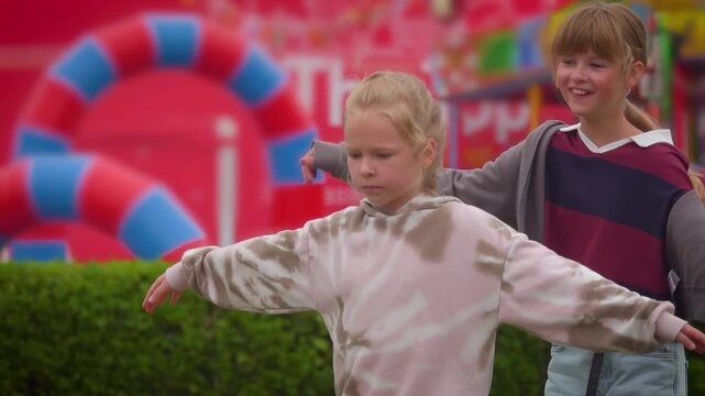 Two carefree young friends walk along a parapet with their arms outstretched. The vibrant, glowing lights of a nighttime amusement park create a magical and dreamy atmosphere