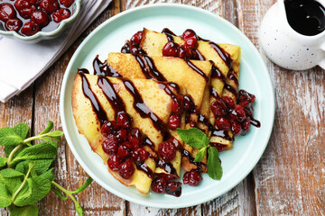 Tasty crepes with cherries, chocolate and mint served on wooden table, flat lay