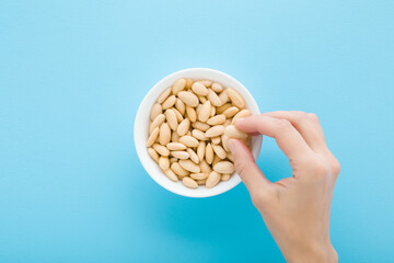 Young adult woman hand fingers taking almond nuts from white bowl on light blue table background. Pastel color. Closeup. Point of view shot. Top down view.