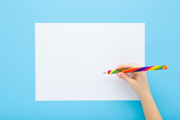 Little child hand holding colorful pencil and drawing on blank white paper on light blue table background. Pastel color. Closeup. Point of view shot. Children creativity time. Top down view.