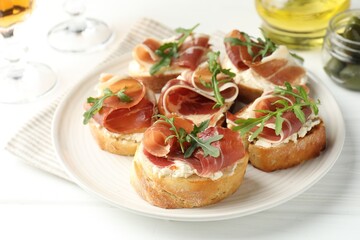 Delicious bruschettas with cream cheese, prosciutto and arugula on white wooden table, closeup