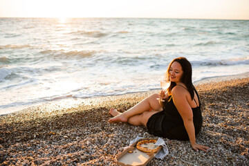 Smiling young woman 35-39 year old holding glass of wine and eating pizza sitting at beach over evening sea outdoors. Summer vacation season.