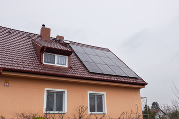 House with orange exterior wall and brown roof featuring solar panels on the sloped surface under cloudy sky with trees in the background and window visible on the side