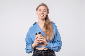 Cheerful young woman holding coffee cup in hands standing against white backdrop wearing casual clothes, modern lifestyle concept