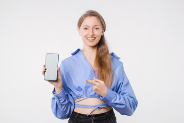 Happy young female showing smartphone while pointing at it, smiling joyfully, studio shot on white background, casual outfit, digital communication concept