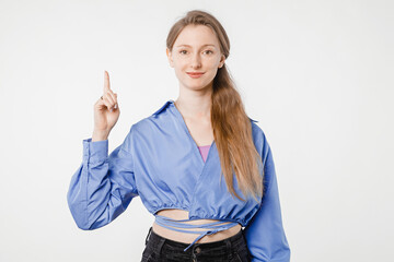 Friendly young woman standing in studio against white background, casually dressed, smiling and pointing with her finger to an empty space for ad, slogan or text, concept of promotion and presentation