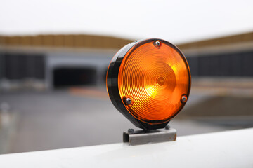 Orange warning light mounted on black bracket with circular lens, illuminated, positioned on white railing with blurred background of road and structures in distance