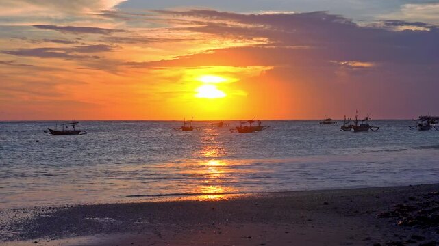 Amazing seascape with setting sun near horizon line, orange sunset sky, silhouettes of traditional fisherman's boats floating in ocean and foamy waves breaking on sand beach. Camera stays still.