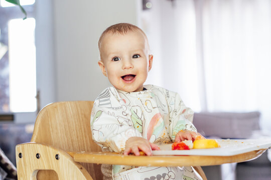 Smiling happy cute baby eating himself on high chair baby led weaning or blw. Infant self-feeding solid food fine motor development. Baby's feeding, learning eating food by himself and making mess