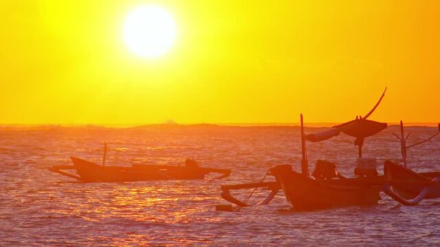 Amazing seascape with setting sun near horizon line, orange sunset sky, silhouettes of traditional fisherman's boats floating in ocean and foamy waves breaking on sand beach. Camera stays still.