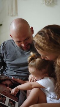 Loving family enjoys time together, watching a photo album on the couch in a cozy, modern home. Concept of parenting, bonding, happy memories of childhood, parenthood. Vertical shot