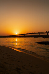Sunset over a sandy beach with a pedestrian bridge silhouetted against a golden sky. Suitable for travel, tourism, and attractions.