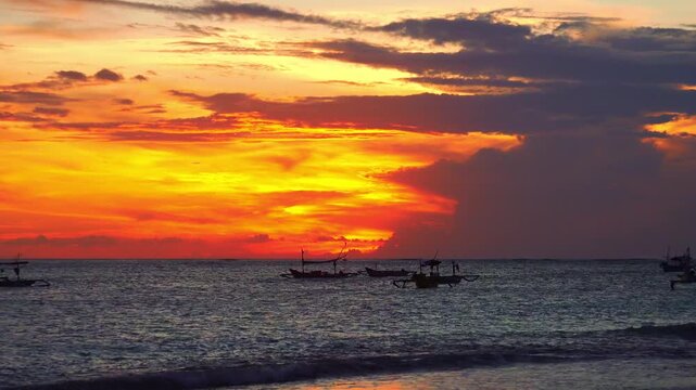 Silhouettes of outrigger fishing boats swaying on sea waves against sun disappearing below horizon. Tropical sunset seascape with ships floating in ocean illuminated by sunlight. Camera zooms out.