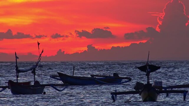 Silhouettes of traditional fishing vessels floating in ocean against red sunset sky with clouds lit by last beams of setting sun on background. Beautiful scene with evening sea. Camera stays still.