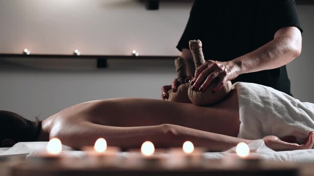 Skilled therapist performs traditional herbal compress massage on a client&rsquo;s back, enhancing relaxation and wellness in a serene candlelit spa. Concept healing, calm, wellness