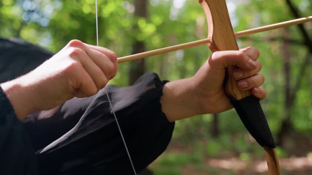 Close up of hands in black attire pulling bowstring with steady focus, sunlight glimmering through green forest, tension captured in graceful motion symbolizing strength