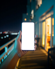 Fototapeta premium First person point of view of a hand holding a smartphone with a blank white screen mockup on a dark outdoor balcony at night with blurred city lights and warm room glow background