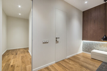 A hallway shows a wooden floor leading to a closed door. The walls are light with a wooden feature panel. Lights on the ceiling brighten the space, creating an inviting atmosphere