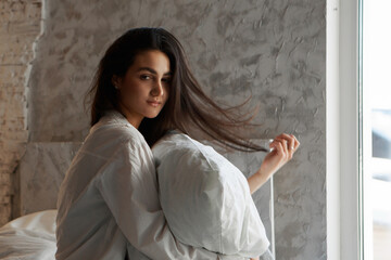 A woman with black hair wakes up in the morning in a white blanket and catches the rays of the sun. The portrait is creative, with brown eyes. High quality photo