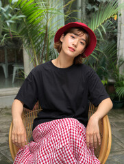 Young woman with short hair red hat black t-shirt and red checkered skirt sitting in a chair.