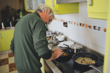 Senior man cooking food in a domestic kitchen, frying fish fillets and croquettes in two pans on an...