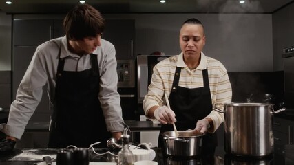 Focused multiethnic female chef stirring sauce in pot demonstrating cooking technique to male trainee during group culinary class in restaurant kitchen