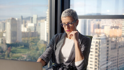 Middle aged businesswoman types on a laptop beside a large window with a panoramic city skyline, focused on daily tasks in a modern corporate office during daylight.