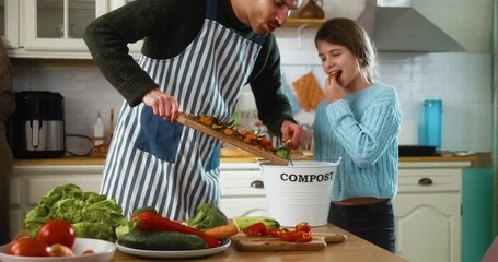 Family throws vegetable leftovers into a compost bin after cooking in the kitchen. Teaching daughter sustainability, recycling food waste, and environmental awareness at home.