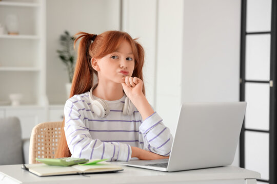 Thoughtful little redhead girl with laptop studying online at home