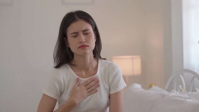 Young woman clutching her chest in pain while sitting on bed, showing signs of heart problem, chest pain, anxiety attack, or breathing difficulty, related to medical emergency symptoms.