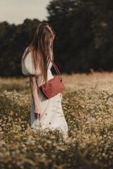 A young woman in a white dress with a red handbag walks among blooming wildflowers in a sunny meadow during the golden hour, captured in an aesthetic vertical orientation