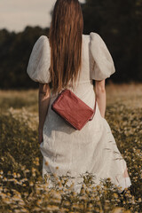 Portrait of a young woman backwards in a white lace dress walking through a blooming chamomile field in the warm light of the setting sun with a red handbag over her shoulder, vertical orientation, ru