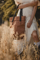 Woman in white lace boho dress holding a stylish brown washpapa bag in an oat field during the golden hour in a vertical frame