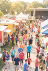High angle blurry bokeh view of a busy outdoor summer festival with crowds of people walking between colorful food trucks and market tents.