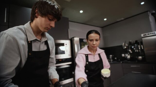 Male chef in apron adding cocoa powder to ramekin with spoon, coating ceramic mold before baking chocolate cupcakes while female trainee learning cooking technique nearby during workshop in kitchen