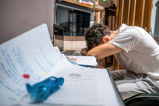 Young man feeling exhausted and overwhelmed, sleeping with head on his desk surrounded by books and laptop after intense studying
