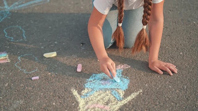 Child draws girl face with chalk on street. Summer outdoor play moment. Girl adds colors with chalk. Art grows on street. Drawing shapes with joy. Kid enjoys sunny day with art and chalk drawing fun.