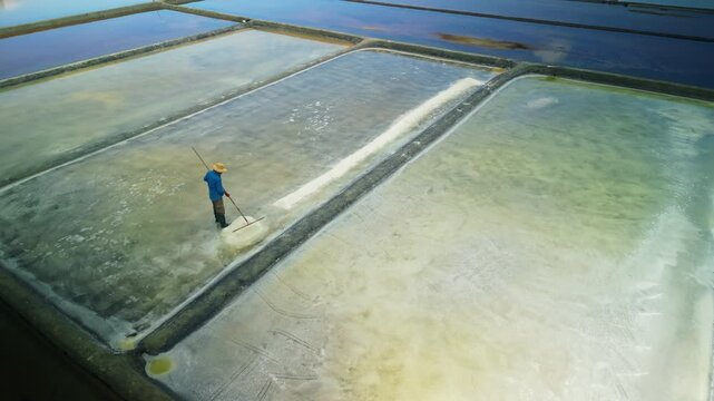 Asian worker collects sea salt crystals using traditional tool in solar evaporation ponds. Aerial view of mineral harvesting process in coastal salt farm and traditional industry.