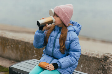 Photograph, portrait of a beautiful teenage girl, child with a thermos of hot tea and croissant, food, sitting on a bench with the sea in the background on a picnic.