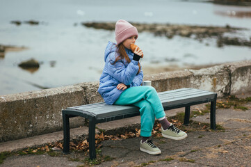 Photograph, portrait of a beautiful teenage girl, child with a thermos and a croissant, sitting on a bench against the backdrop of the sea on a picnic.
