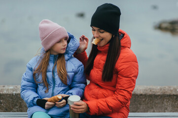 Photo, portrait of teenage girl, child with thermos of hot tea and croissant, food, sitting with mother, young woman on bench against sea background on picnic in autumn.