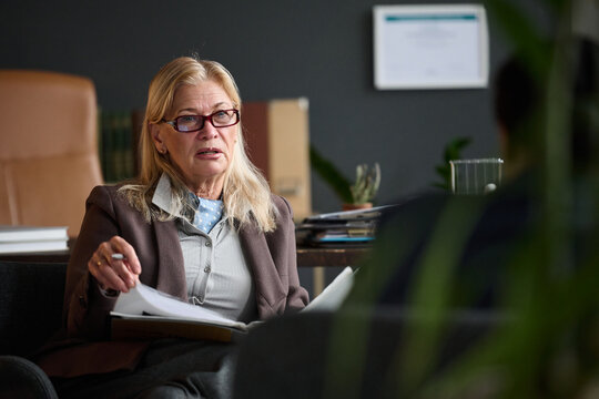 Caucasian senior woman therapist conducting counseling session with unseen client in office setting, holding documents and gesturing while listening attentively during therapy