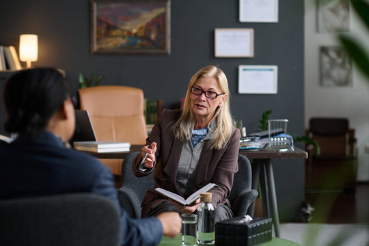Senior Caucasian therapist holding notebook talking to young adult Black woman during therapy session in office setting, both seated facing each other, water glasses on table