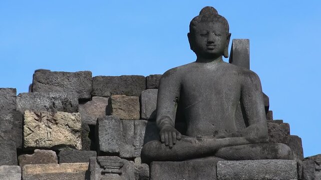 Statue of meditating Buddha carved from dark stone against clear blue sky on background, decoration of ancient Borobudur Mahayana Buddhist temple in Magelang, Central Java, Indonesia. Panning video.