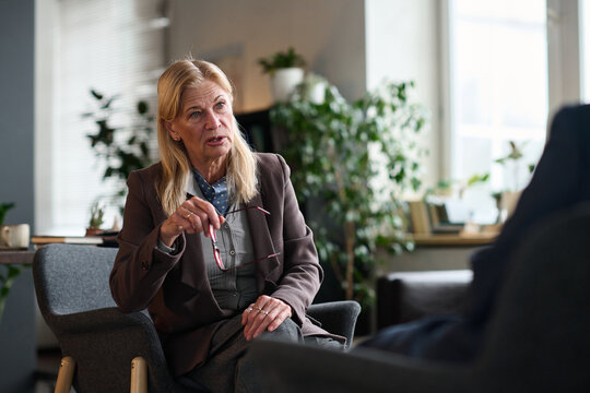 Senior Caucasian woman sitting in therapy session holding eyeglasses, listening attentively to unseen client, engaging in professional counseling conversation in office setting