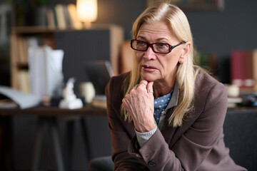 Caucasian senior woman wearing glasses sitting in office setting listening attentively during therapy session, hand resting on chin, appearing focused and engaged in conversation