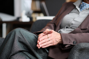 Senior Caucasian woman sitting with hands clasped during therapy session, wearing business attire, showing attentive posture, partial body visible, focus on hands and lap