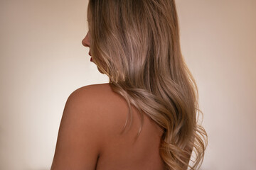Rear view of young blonde woman with long wavy blonde hair and bare shoulders on neutral...